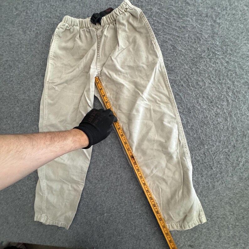K&ouml;nnte beinhalten: Beigefarbene Hose mit elastischem Bund und integriertem schwarzen Schnalleng&uuml;rtel. Ein gelbes Ma&szlig;band liegt entlang der inneren Beinnaht und zeigt eine L&auml;nge von etwa 61 cm vom Schritt bis zum Saum an.