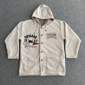Op de afbeelding: Lichtgrijze jas met capuchon, knoopsluiting en twee voorzakken. De jas heeft de tekst "CHICAGO 1967 BRANSON" en "THE BRAND WITH THE 3 STRIPES" in donkergroen en kastanjebruin. De capuchon is gevoerd met een bruine stof.