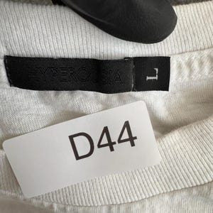 May include: Close-up of a white t-shirt with a black label that reads "HYPEROSIA" and a size "L". A white tag with the number "D44" is attached to the shirt.