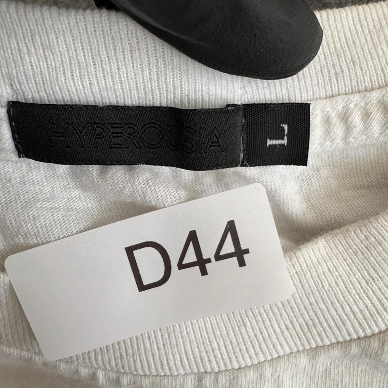 May include: Close-up of a white t-shirt with a black label that reads "HYPEROSIA" and a size "L". A white tag with the number "D44" is attached to the shirt.