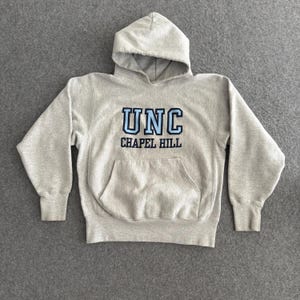 May include: A light gray hooded sweatshirt with the text "UNC CHAPEL HILL" in blue and black. The sweatshirt has a front pocket and ribbed cuffs and hem. The hood is unlined.
