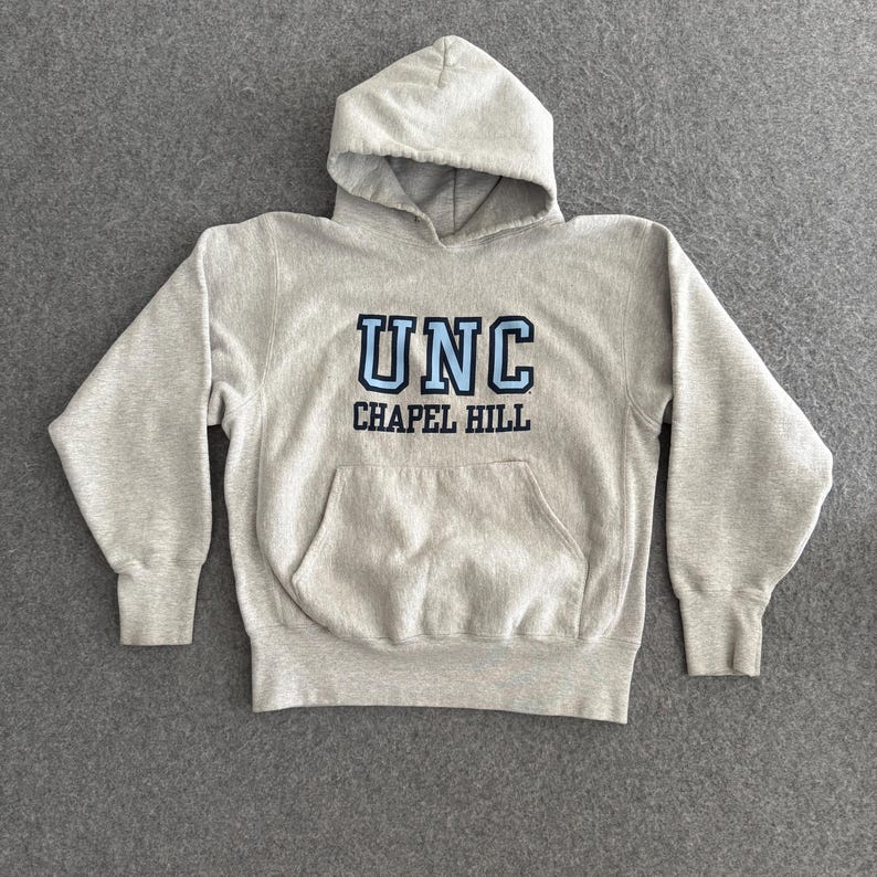 May include: A light gray hooded sweatshirt with the text "UNC CHAPEL HILL" in blue and black. The sweatshirt has a front pocket and ribbed cuffs and hem. The hood is unlined.