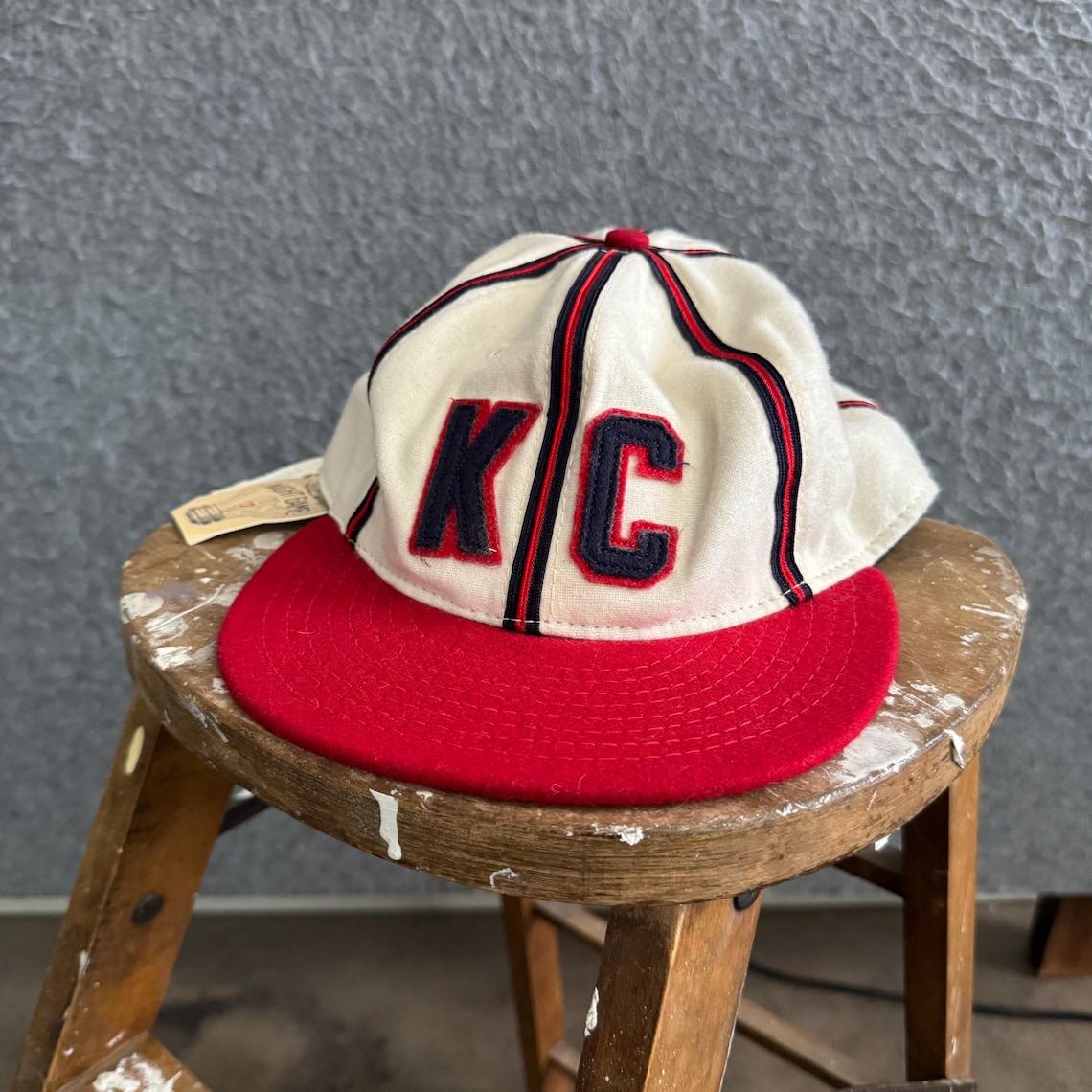 May include: A cream-colored baseball cap featuring a red brim and dark blue embroidered letters KC on the front. The cap has red and dark blue vertical piping along the seams of the crown and includes a small paper tag attached to the side.