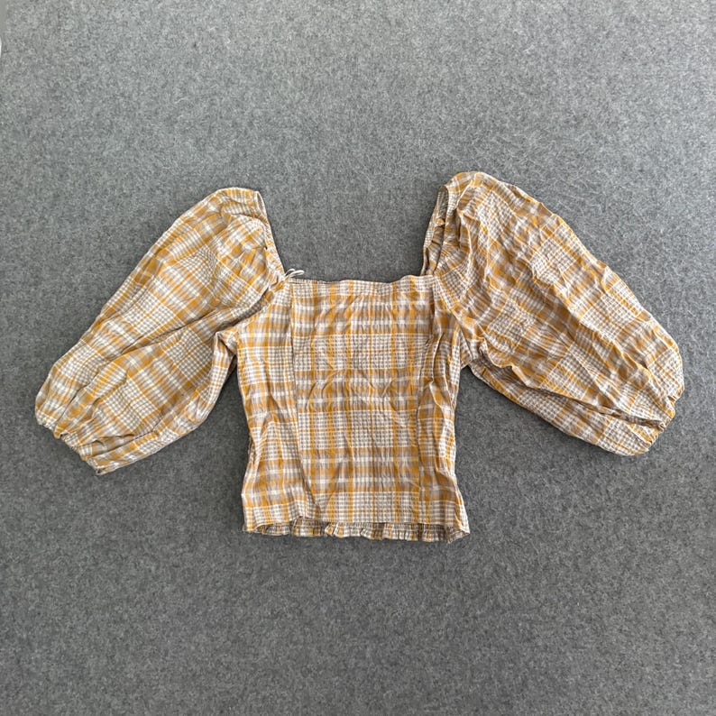 May include: A yellow and white checked top with a square neckline and long, puffed sleeves. The top is made of a lightweight fabric and is laid flat on a grey surface. The top is a casual style.