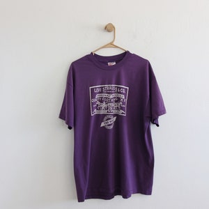 Puede incluir: Una camiseta morada con un gráfico blanco con el texto "LEVI STRAUSS & CO. ORIGINAL RIVETED QUALITY CLOTHING". La camiseta está colgada en una percha de madera contra una pared blanca.