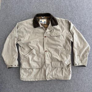 May include: A tan jacket with a brown corduroy collar and a plaid lining. The jacket has four front pockets, button closures, and a small logo on the chest. The jacket is laid flat on a gray surface.