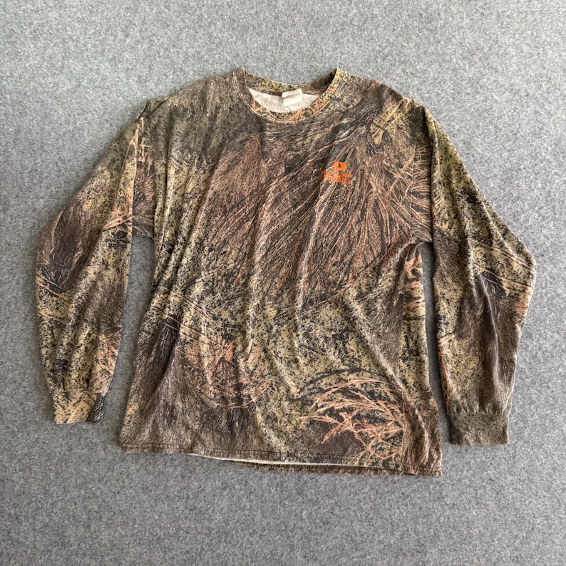 May include: Long-sleeved camouflage shirt in shades of brown, green, and tan. The shirt has a crew neck and a small orange logo on the chest. The fabric appears to be lightweight.