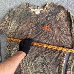 May include: Camouflage long-sleeve shirt with the Mossy Oak logo in orange. The shirt is brown, tan, and green. A measuring tape is held across the shirt. The shirt is made of a soft, comfortable material.