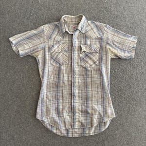 May include: A short-sleeved, button-down shirt with a plaid pattern in shades of cream, blue, and brown. The shirt has two chest pockets and a classic collar. The shirt is made of a lightweight fabric.