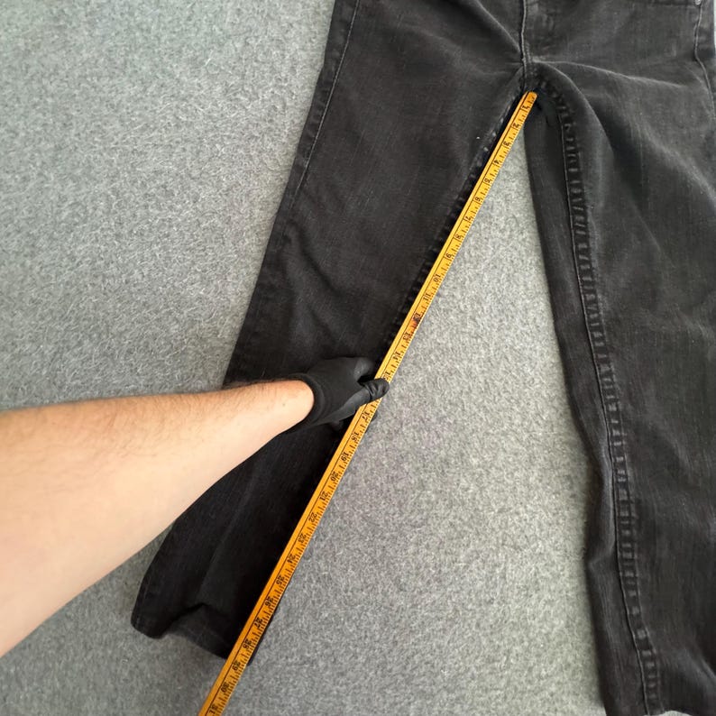 Pu&ograve; includere: Jeans neri in denim con design a gamba dritta. Un righello di legno giallo viene utilizzato per misurare la lunghezza dei jeans. I jeans sono appoggiati su una superficie grigia.