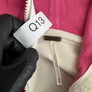 Op de afbeelding: Close-up van een roze en cr&egrave;mekleurige hoodie met ritssluiting. De hoodie heeft een witte rits en een label met de tekst "Free People". Het label heeft ook de tekst "MADE IN CHINA FABRIQUE EN CHINE". Een wit label met "Q13" is ook zichtbaar.