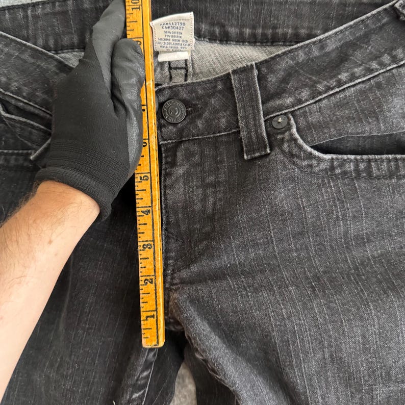 Pu&ograve; includere: Primo piano di jeans neri con chiusura a bottone e passanti per cintura. Un righello giallo &egrave; tenuto contro i jeans, mostrando le misure in centimetri. I jeans hanno un aspetto sbiadito e usurato.