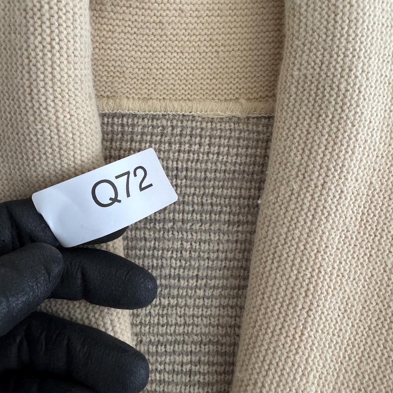 May include: A beige knit cardigan featuring a thick ribbed collar and a textured weave pattern. A white rectangular tag with the black printed text Q72 is attached to the garment, showing the knit construction and the contrast between the collar.