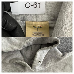 May include: Close-up of a gray hoodie with a drawstring hood. The tag reads "The Game" and "S". The hoodie is made of 90% cotton and 10% polyester. The tag also includes washing instructions and "MADE IN THE USA".