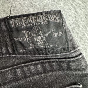 Pu&ograve; includere: Primo piano di una toppa in denim nero dei jeans True Religion. La toppa presenta il nome del marchio, una grafica di una figura che suona la chitarra e le parole "World Tour".
