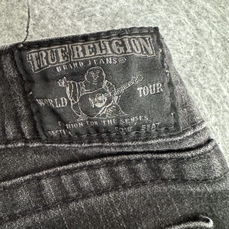 Pu&ograve; includere: Primo piano di una toppa in denim nero dei jeans True Religion. La toppa presenta il nome del marchio, una grafica di una figura che suona la chitarra e le parole "World Tour".