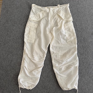 May include: Off-white cargo pants with multiple pockets and drawstring cuffs. The pants have a high waist and a relaxed fit. The fabric appears lightweight and slightly wrinkled. The pants are laid flat on a gray surface.