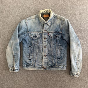 May include: Light blue denim jacket with a button-up front, two chest pockets, and two side pockets. The jacket has a classic collar and long sleeves with buttoned cuffs. The denim has a faded, worn look, with a distressed collar.