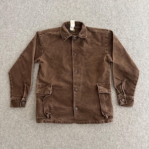 May include: A brown long-sleeve button-up jacket featuring a standard collar, five front buttons, and two large patch pockets with flaps at the lower front. The garment has buttoned cuffs and a visible brand tag at the inner neckline.