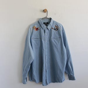 Puede incluir: Una camisa vaquera azul claro con personajes de dibujos animados bordados en los hombros. Los personajes son Bugs Bunny y Yosemite Sam, ambos con sombreros de vaquero y sosteniendo cuerdas.