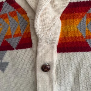 May include: A cream-colored knit cardigan featuring a shawl collar and a single dark brown button. The garment displays a horizontal pattern of red, orange, and yellow stripes accented with gray geometric shapes across the chest area.