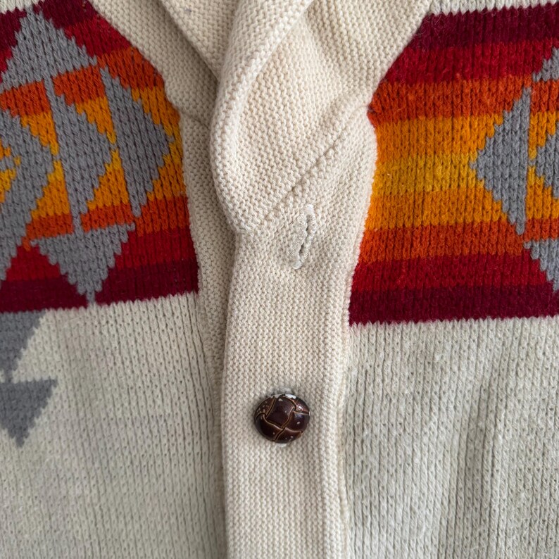 May include: A cream-colored knit cardigan featuring a shawl collar and a single dark brown button. The garment displays a horizontal pattern of red, orange, and yellow stripes accented with gray geometric shapes across the chest area.
