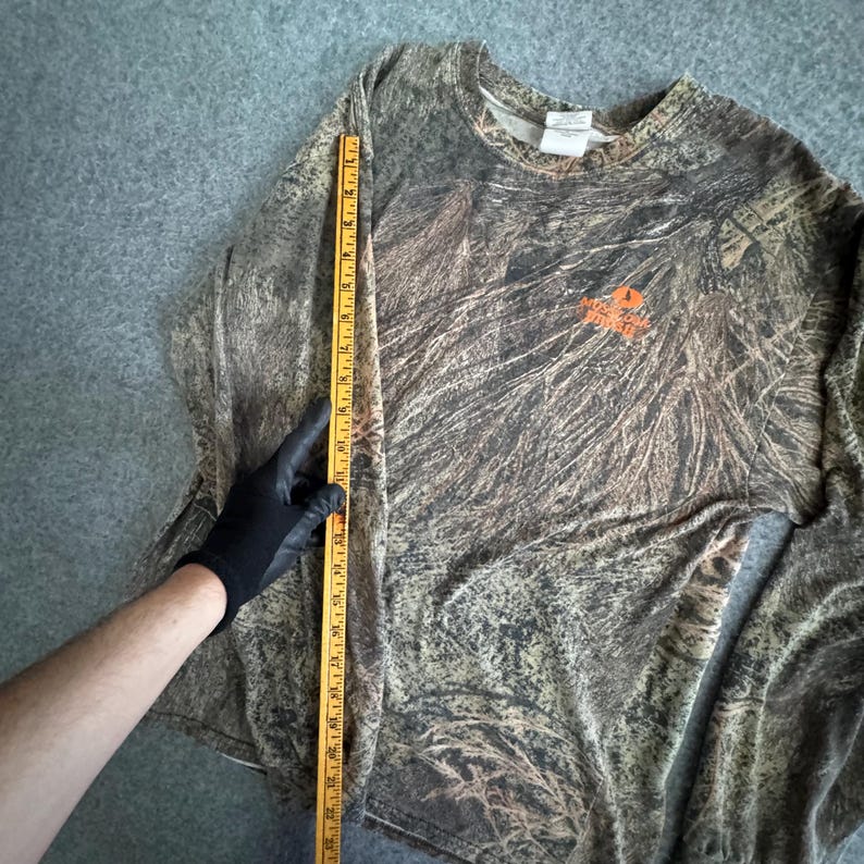 May include: Camouflage long-sleeve shirt with the Mossy Oak logo in orange. The shirt is measured with a yellow ruler. The shirt has a pattern of brown, green, and tan colors.