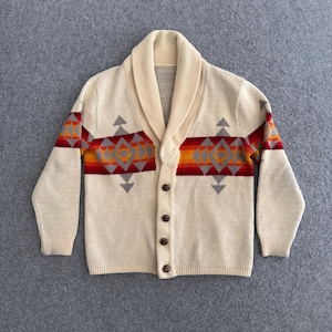 May include: A cream-colored knit cardigan featuring a shawl collar and a front button closure. The garment displays a horizontal band across the chest and sleeves with a geometric pattern in shades of orange, red, and grey against the cream base.