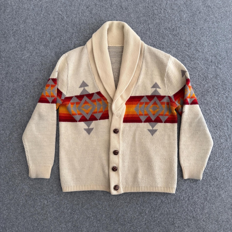 May include: A cream-colored knit cardigan featuring a shawl collar and a front button closure. The garment displays a horizontal band across the chest and sleeves with a geometric pattern in shades of orange, red, and grey against the cream base.