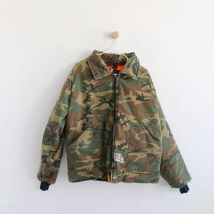 Puede incluir: Una chaqueta de camuflaje con cierre de cremallera y forro negro. La chaqueta tiene un bolsillo en el lado izquierdo y un pequeño bolsillo en el lado derecho. La chaqueta está colgada en una percha blanca contra una pared blanca.