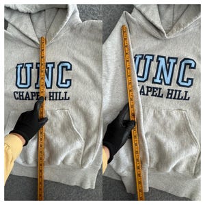 May include: A light gray hoodie with a front pocket and a hood. The hoodie features the text "UNC CHAPEL HILL" in blue and white. A measuring tape is held up to the hoodie.
