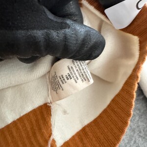 Op de afbeelding: Close-up van een gebreide trui met afwisselende strepen in wit en een warme bruine kleur. Een label met tekst in meerdere talen, waaronder "HECHO EN CHINA" en "FABRICADO NA CHINA", is zichtbaar. De trui wordt gedeeltelijk bedekt door een zwarte handschoen.