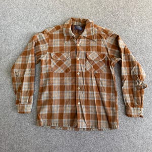 Puede incluir: Camisa de manga larga con estampado de cuadros en marrón, beige y azul claro. Presenta cierre de botones, dos bolsillos en el pecho y cuello clásico. La tela parece ser de peso medio.