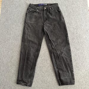 May include: Faded black denim jeans with a relaxed fit. The jeans feature a classic five-pocket design, a button and zip closure, and the word "BAGGY" is visible on the waistband. The jeans are laid flat on a grey surface.