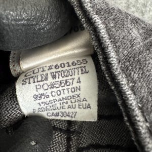 Pu&ograve; includere: Primo piano di un'etichetta di abbigliamento con testo. L'etichetta recita "CUT#601655 STYLEN W70207TEL PO#56574 99% COTTON 1% SPANDEX MADE IN USA FABRIQUE AU EUA CA#30427". L'etichetta &egrave; bianca con testo nero ed &egrave; attaccata a tessuto grigio.