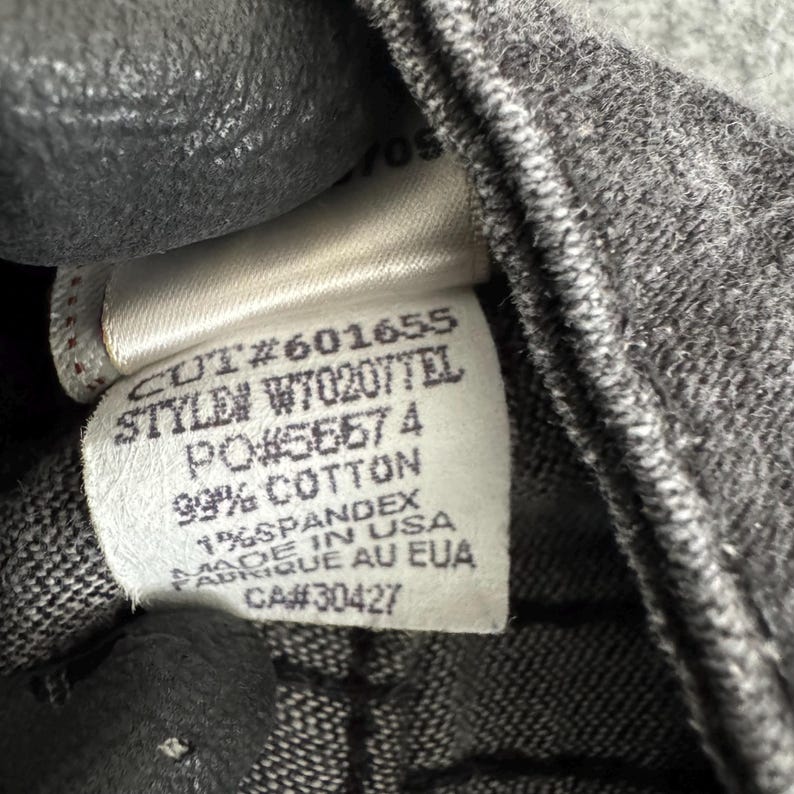 Pu&ograve; includere: Primo piano di un'etichetta di abbigliamento con testo. L'etichetta recita "CUT#601655 STYLEN W70207TEL PO#56574 99% COTTON 1% SPANDEX MADE IN USA FABRIQUE AU EUA CA#30427". L'etichetta &egrave; bianca con testo nero ed &egrave; attaccata a tessuto grigio.