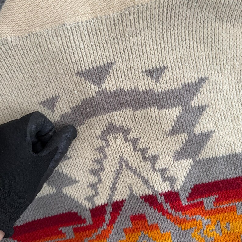 May include: A beige knit sweater featuring a geometric pattern in shades of gray, red, and orange. The knit texture is visible throughout the garment, showcasing a repeating triangular and stepped design across the front surface of the fabric.