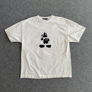 May include: Off-white t-shirt featuring a black and white graphic of a cartoon mouse. The short-sleeved shirt has a crew neck and a relaxed fit. The mouse graphic is centered on the front of the shirt.