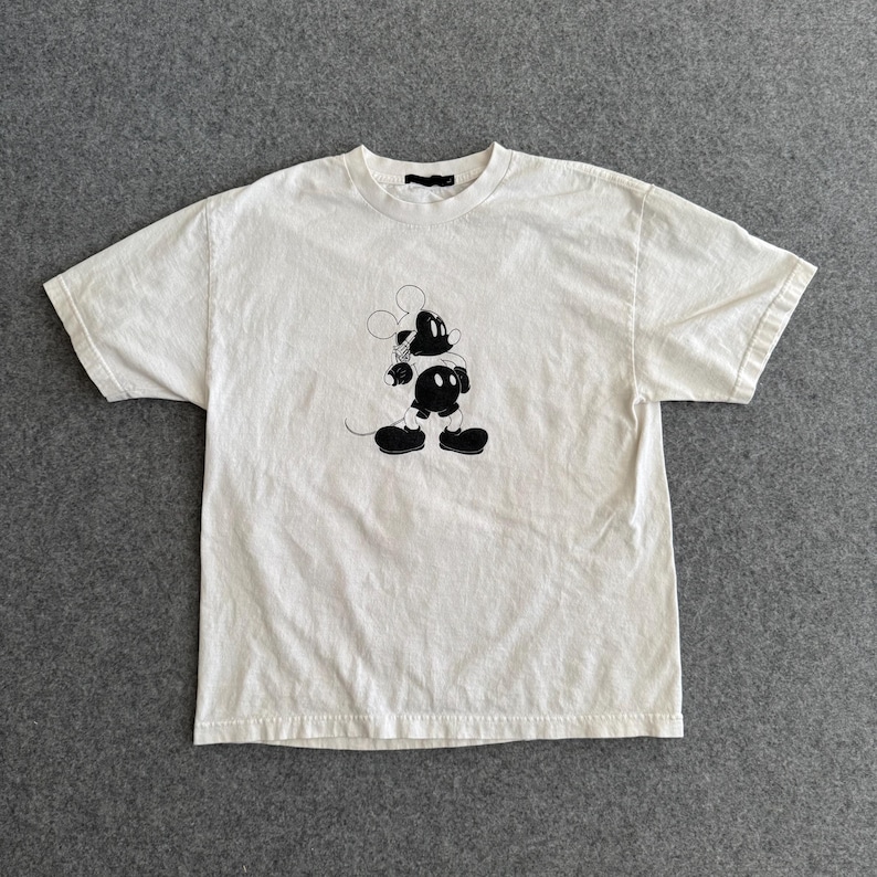 May include: Off-white t-shirt featuring a black and white graphic of a cartoon mouse. The short-sleeved shirt has a crew neck and a relaxed fit. The mouse graphic is centered on the front of the shirt.