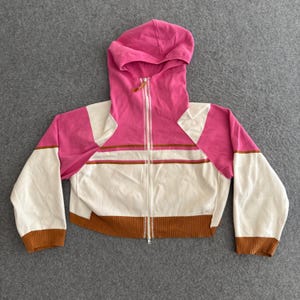 Op de afbeelding: Een cropped zip-up hoodie in een colorblock design. De hoodie heeft een roze capuchon en schouderpanelen, witte mouwen en body en bruine accenten. De jas heeft een volledige rits en bruine geribde manchetten.