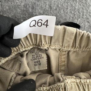 K&ouml;nnte beinhalten: Eine beigefarbene Hose mit elastischem Bund und einem wei&szlig;en Etikett mit der Aufschrift Q64. Das Innenetikett zeigt Made in U.S.A., RN 76104, CA 25124 und Kids 10/12. Der Stoff ist hellbeige mit sichtbaren N&auml;hten am Hosenbund.