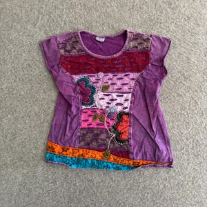 May include: A short-sleeved purple t-shirt featuring horizontal patchwork panels in shades of red, pink, and lavender. The garment includes decorative floral appliqués, embroidered details, and a bottom border with orange and blue floral patterns.