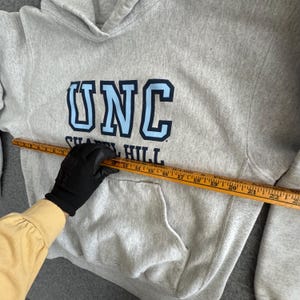 May include: A light gray hooded sweatshirt with a front pocket and the letters "UNC" in light blue and navy blue. The text "Chapel Hill" is below the letters. A wooden ruler is placed across the sweatshirt.