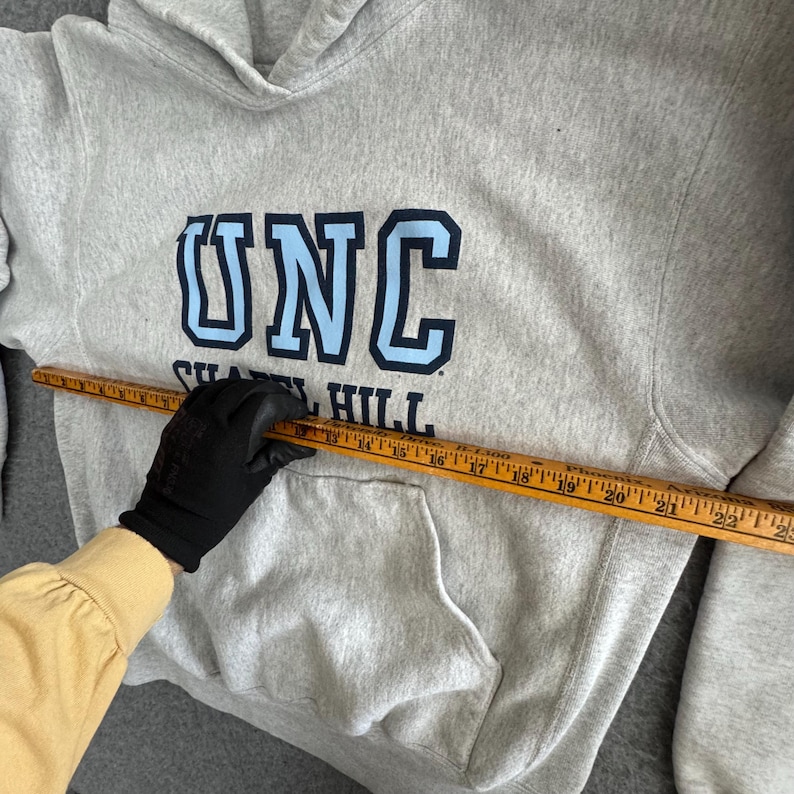May include: A light gray hooded sweatshirt with a front pocket and the letters "UNC" in light blue and navy blue. The text "Chapel Hill" is below the letters. A wooden ruler is placed across the sweatshirt.