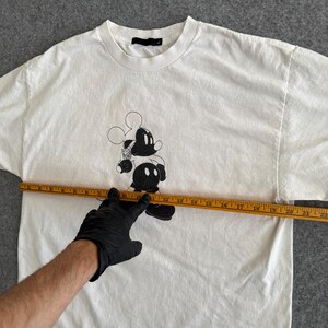 May include: White t-shirt featuring a black and white graphic of Mickey Mouse. The shirt has a crew neck and short sleeves. A measuring tape is placed across the shirt.