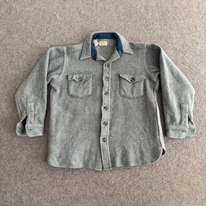 May include: A gray button-down shirt with two chest pockets and a navy blue collar. The shirt has long sleeves with buttoned cuffs. The fabric has a textured, herringbone pattern. The shirt has a small tag on the collar.