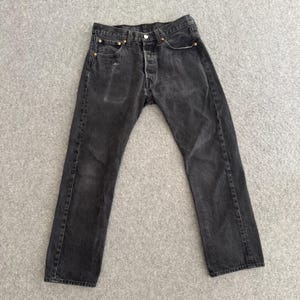 May include: A pair of faded black denim jeans. The jeans have a button fly, classic five-pocket design, and copper rivets. The fabric shows signs of wear and fading, giving them a vintage look. The jeans are laid flat on a gray surface.
