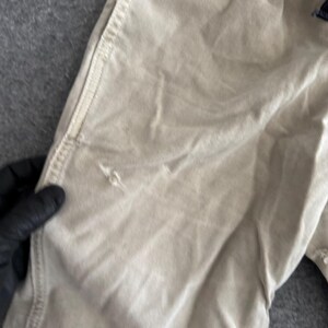 K&ouml;nnte beinhalten: Eine beigefarbene Hose mit einer strukturierten Stoffkonstruktion, sichtbaren vertikalen N&auml;hten entlang der Seitennaht und einem kleinen, deutlichen Riss oder Faden im Material auf der Vorderseite, der die Webart des Kleidungsst&uuml;cks zeigt.