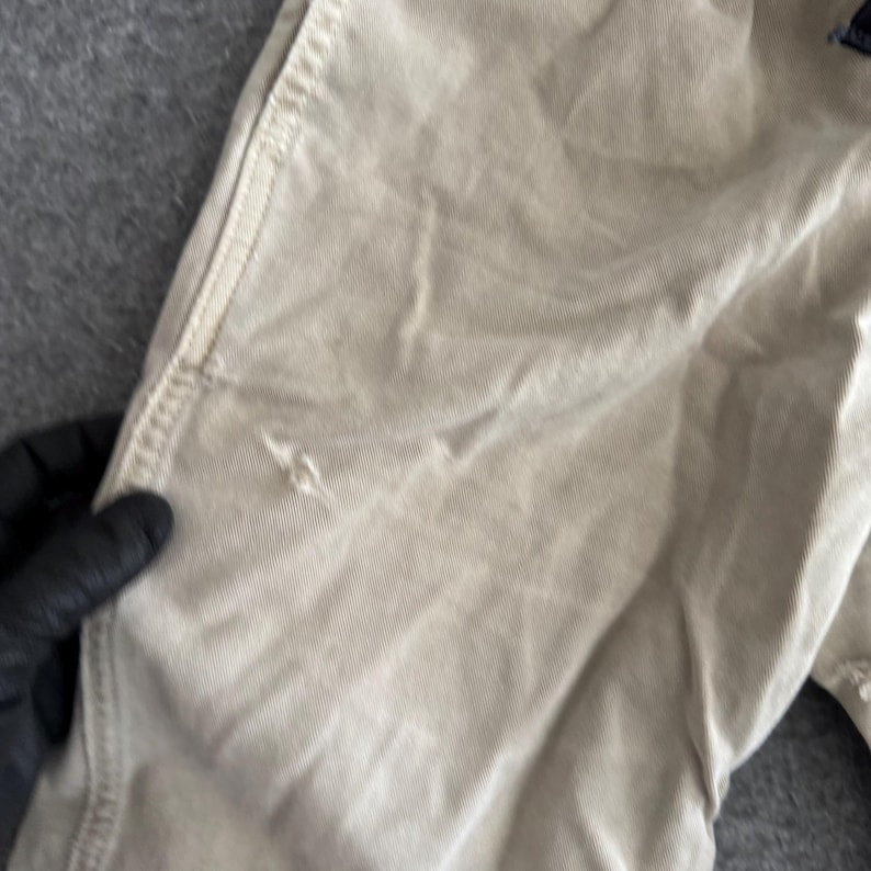 K&ouml;nnte beinhalten: Eine beigefarbene Hose mit einer strukturierten Stoffkonstruktion, sichtbaren vertikalen N&auml;hten entlang der Seitennaht und einem kleinen, deutlichen Riss oder Faden im Material auf der Vorderseite, der die Webart des Kleidungsst&uuml;cks zeigt.