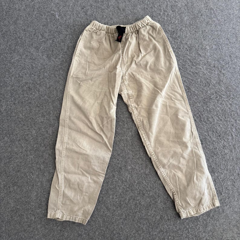 K&ouml;nnte beinhalten: Beigefarbene Baumwollhose mit elastischem Bund und einem schwarzen Innenetikett. Die Hose hat eine lockere Passform mit sichtbaren vertikalen Nahtdetails an der Vorderseite jedes Beins und eine leicht zulaufende Silhouette an den Kn&ouml;cheln.